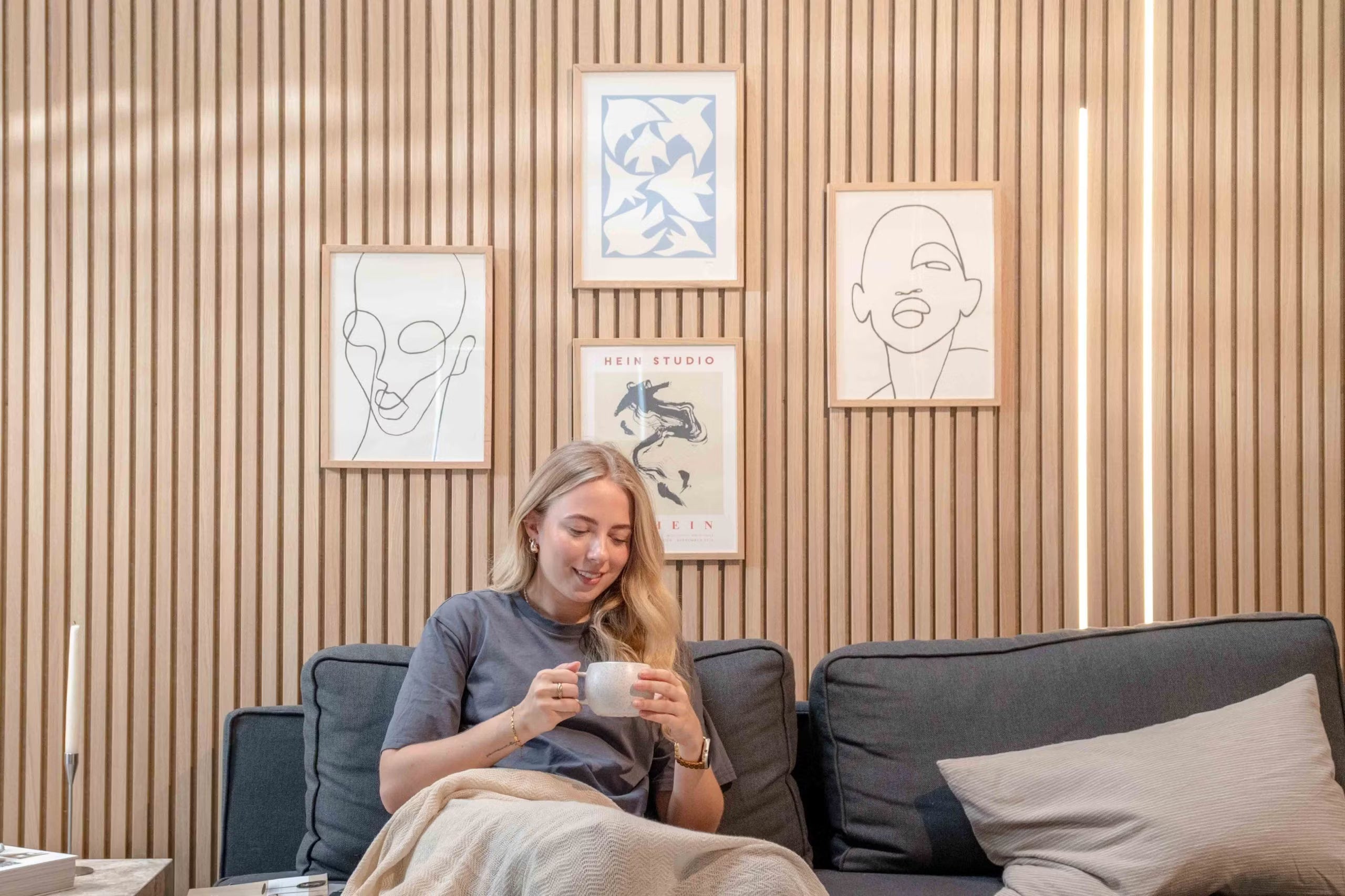 Femme se relaxant sur un canapé avec une tasse, souriant sous un tableau minimaliste et un décor abstrait montés sur des panneaux acoustiques texturés en lattes nordiques.